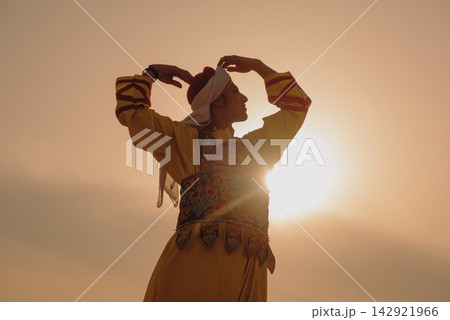 Traditional folk dancer in embroidered Egyptian heritage costume performing arms raised pose against warm golden sunset sky with dramatic lens flare Traditional folk dancer in embroidered Egyptian heritage costume performing arms raised pose against warm golden sunset sky with dramatic lens flare 142921966