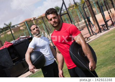 Middle Eastern men working out together in outdoor CrossFit gym holding medicine ball and weight plate Middle Eastern men working out together in outdoor CrossFit gym holding medicine ball and weight plate 142922288