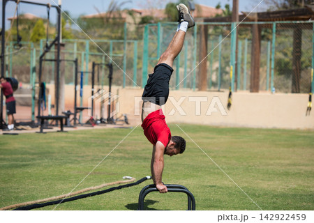 Man performing handstand on parallettes at outdoor crossfit gym with friend nearby Man performing handstand on parallettes at outdoor crossfit gym with friend nearby 142922459