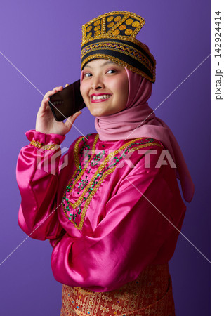 Smiling Young Woman in Hijab and Traditional Southeast Asian Headdress Talking on Smartphone Smiling Young Woman in Hijab and Traditional Southeast Asian Headdress Talking on Smartphone 142924414