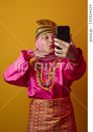 Young Woman in Traditional Southeast Asian Costume and Hijab Taking a Colorful Selfie With Phone Young Woman in Traditional Southeast Asian Costume and Hijab Taking a Colorful Selfie With Phone 142924424