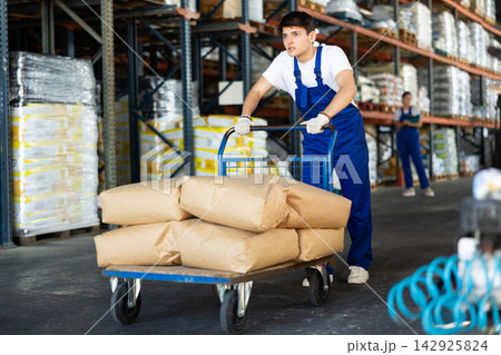 In warehouse of store, stevedore loader young man pushes and carries large cart for bulky cargo In warehouse of store, stevedore loader young man pushes and carries large cart for bulky cargo 142925824