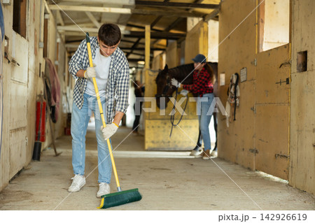 Young male worker cleaning floor at stabling, woman farmer standing with horse in background 142926619
