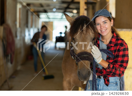 European young girl rancher holding rein while standing with horse in stable and petting horse, looking at camera. Woman cleaning floor in background 142926630