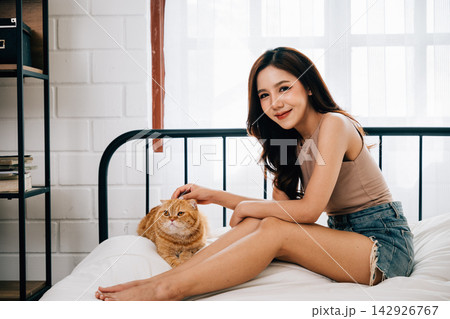 In a portrait of comfort and togetherness, a happy young woman and her Scottish Fold cat enjoy resting in bed at home. Their friendship is evident through joyful embraces and content smiles. In a portrait of comfort and togetherness, a happy young woman and her Scottish Fold cat enjoy resting in bed at home. Their friendship is evident through joyful embraces and content smiles. 142926767