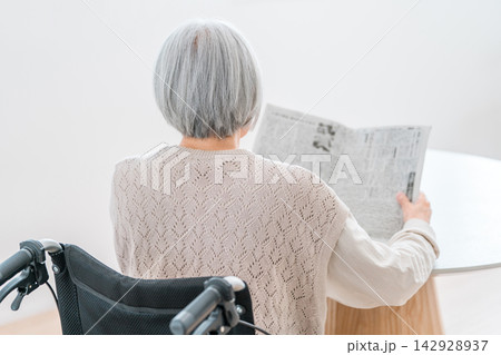 車椅子に座って新聞を読むグレイヘアのシニア女性の後ろ姿。老後の情報収集 車椅子に座って新聞を読むグレイヘアのシニア女性の後ろ姿。老後の情報収集 142928937