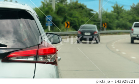 Rear view white car driving along a wide open concrete road. Curved sloping road. A car is overtaking and passing ahead. Side road with guardrail and green trees under blue sky. 142929221