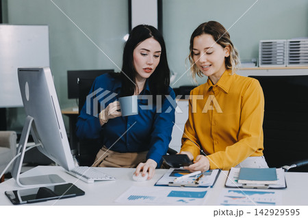 Two businesswomen discussing work using a laptop in a modern office lounge area, showing teamwork and communication 142929595