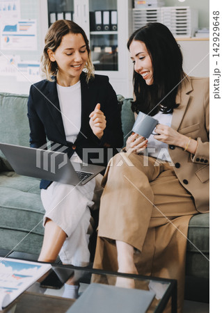 Two smiling women are comfortably sitting on a couch, sharing a moment and collaborating on a tablet, working together in a home office environment 142929648