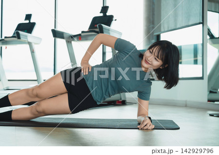 Young woman performing side plank exercise on mat in modern gym with treadmill equipment and large windows Young woman performing side plank exercise on mat in modern gym with treadmill equipment and large windows 142929796