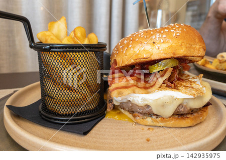 Gourmet hamburger with bacon, cheese, and pickles served with crispy fries in a restaurant setting 142935975