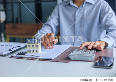 Businessman using calculator and reading contract next to miniature house on the table. Concept for real estate investment, loan, rent, mortgage, insurance Businessman using calculator and reading contract next to miniature house on the table. Concept for real estate investment, loan, rent, mortgage, insurance 142936196