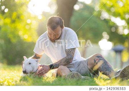 Old bull terrier sits on grass while male owner with tattoos gently pets the dog in a sunlit park setting surrounded by trees and soft greenery Old bull terrier sits on grass while male owner with tattoos gently pets the dog in a sunlit park setting surrounded by trees and soft greenery 142936383