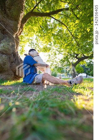Old bull terrier dog sitting on grass with owner in blue shirt, enjoying a sunny day under a large tree in a park setting with green foliage and soft sunlight Old bull terrier dog sitting on grass with owner in blue shirt, enjoying a sunny day under a large tree in a park setting with green foliage and soft sunlight 142936405