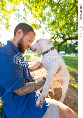 Old bull terrier interacts with male owner in a park, both seated on the ground surrounded by green grass and trees under bright sunlight Old bull terrier interacts with male owner in a park, both seated on the ground surrounded by green grass and trees under bright sunlight 142936407