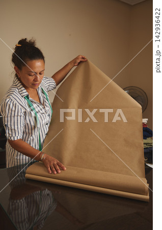 Woman wrapping brown paper roll in a sewing workshop with measuring tape and focused expression Woman wrapping brown paper roll in a sewing workshop with measuring tape and focused expression 142936422