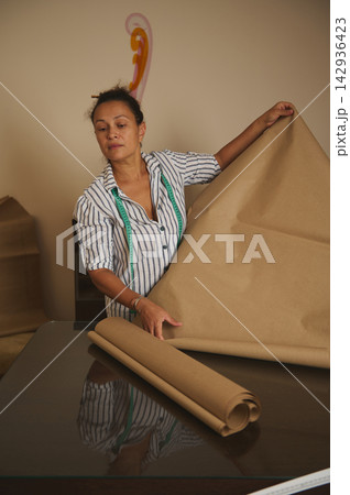 Woman unrolling kraft paper on table with measuring tape preparing patterns for sewing and crafting Woman unrolling kraft paper on table with measuring tape preparing patterns for sewing and crafting 142936423