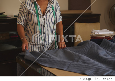 Woman tailor measuring and arranging blue fabric on table with green measuring tape Woman tailor measuring and arranging blue fabric on table with green measuring tape 142936427