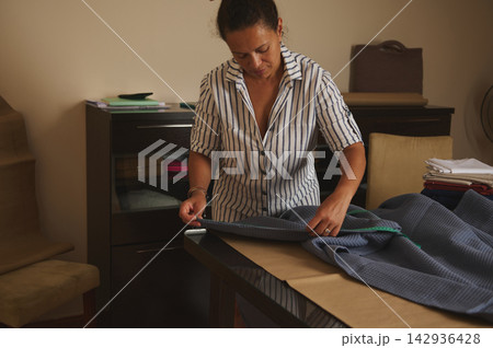 Woman measuring and cutting fabric at a home tailoring workshop preparing garments 142936428