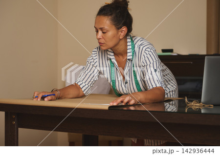 Woman measuring and marking a paper pattern on a table in a home sewing workshop 142936444