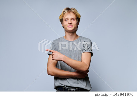 Smiling teenage boy pointing finger to side looking at camera on grey background 142941976