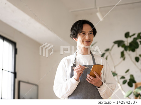 エプロン姿の男性がメモを取りながら考え事をしている様子 142942537