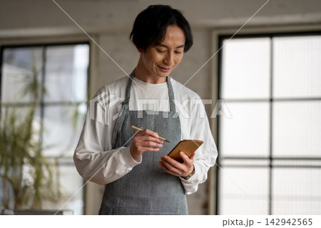 エプロン姿の男性がメモ帳に書き込みながら笑顔で仕事をしている様子 エプロン姿の男性がメモ帳に書き込みながら笑顔で仕事をしている様子 142942565