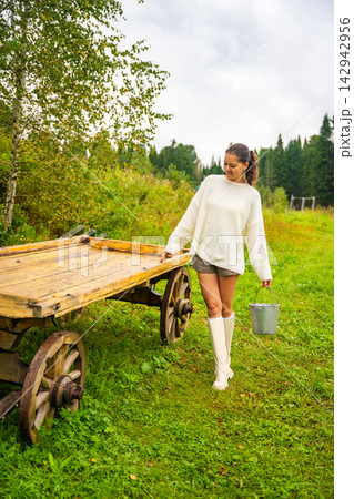 Young woman in white sweater and shorts walks beside wooden cart on grassy field, carrying a bucket, surrounded by trees and cloudy sky in rural landscape 142942956