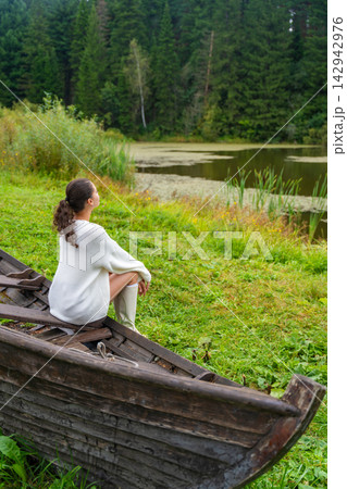 Young woman in white sweater and knee-high boots sits on an old wooden boat, surrounded by lush green grass and trees in a natural outdoor setting 142942976