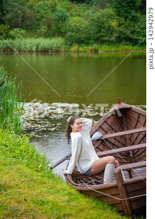 Young woman relaxing in wooden rowboat by lake on autumn day. Cozy sweater and rubber boots represent seasonal outdoor lifestyle and slow living in nature 142942979