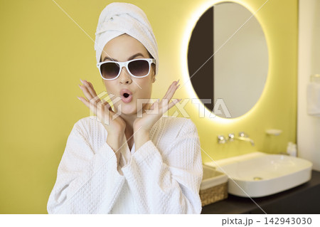 Playful woman in bathrobe and towel with sunglasses in bright modern bathroom 142943030