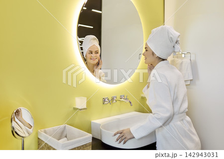 Woman in robe touching face at bathroom mirror, skincare and wellness daily 142943031