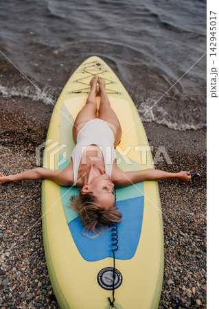 Relaxed woman lying on paddleboard by rocky shoreline wearing white swimsuit during outdoor leisure activity Relaxed woman lying on paddleboard by rocky shoreline wearing white swimsuit during outdoor leisure activity 142945017