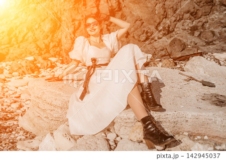 Woman beach fashion sits on rugged coastal rocks wearing a white vintage dress and boots during a golden sunset 142945037