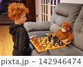 A red hair boy is playing a game of chess with a stuffed red hair monkey. 142946474
