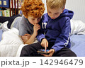 Two children lie on a bed, focused on a smartphone 142946479