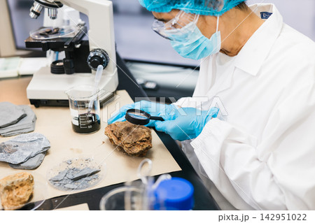 Laboratory researcher in protective gear examines a rock sample with a magnifying glass beside a microscope and beakers. Laboratory researcher in protective gear examines a rock sample with a magnifying glass beside a microscope and beakers. 142951022