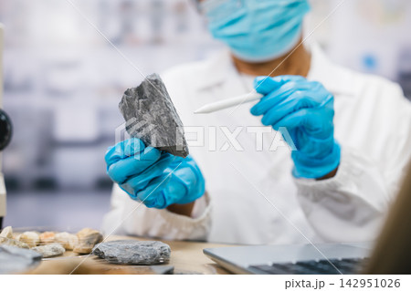 Geologist in a mask and blue gloves examines a rock sample with a probe in a laboratory, with mineral specimens and tools on the worktable. Geologist in a mask and blue gloves examines a rock sample with a probe in a laboratory, with mineral specimens and tools on the worktable. 142951026
