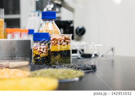 Glass jars of colorful beans, lentils and seeds on a laboratory table beside safety glasses and research equipment. 142951032