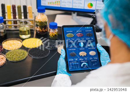 Food scientist in protective clothing uses a tablet to review grain data in a laboratory with samples of beans, seeds, rice, corn and cereals. Food scientist in protective clothing uses a tablet to review grain data in a laboratory with samples of beans, seeds, rice, corn and cereals. 142951048