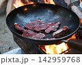 焚き火の上でスキレットに肉を焼くキャンプアウトドア料理 142959768