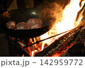 焚き火の炎の上でスキレットに肉を焼くキャンプ料理シーン 142959772
