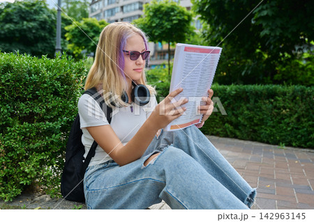 Teenage girl wearing sunglasses reading book sitting on pavement in park Teenage girl wearing sunglasses reading book sitting on pavement in park 142963145