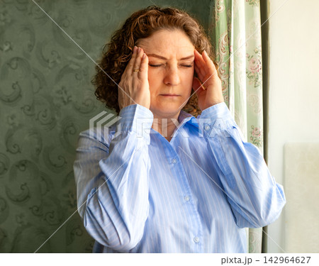 Middle-aged woman holding her head in pain at home, portrait showing headache and stress symptoms Middle-aged woman holding her head in pain at home, portrait showing headache and stress symptoms 142964627