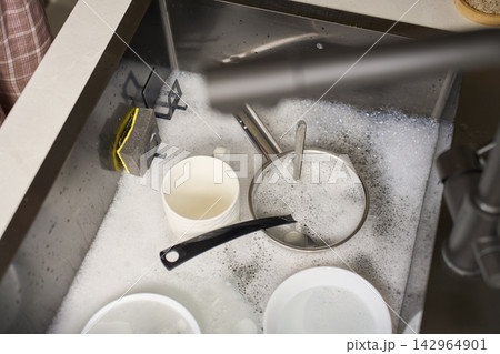 Dishes, saucepan and mug sit in foamy water with cleaning sponge inside kitchen sink 142964901