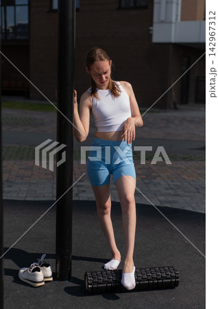 A Caucasian woman stretches her feet with a foam roller at a sports ground. A Caucasian woman stretches her feet with a foam roller at a sports ground. 142967312