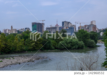 Scenic landscape photography of a wide river and a developing urban skyline with high-rises and construction cranes under an overcast sky Scenic landscape photography of a wide river and a developing urban skyline with high-rises and construction cranes under an overcast sky 142968532
