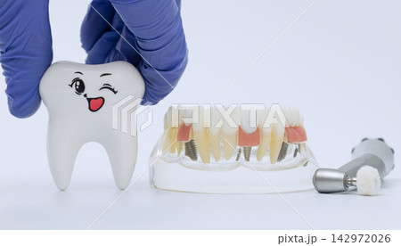 Overcoming dental fear concept with doctor hand holding a funny smiling tooth character near a clear acrylic prosthesis model and medical handpiece. 142972026