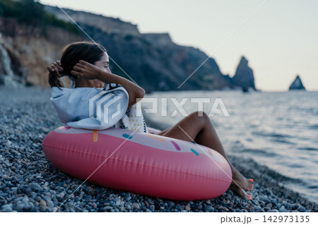 Woman, beach, donut float. Young woman relaxes on pebble beach with inflatable donut by sea, copy space. Woman, beach, donut float. Young woman relaxes on pebble beach with inflatable donut by sea, copy space. 142973135