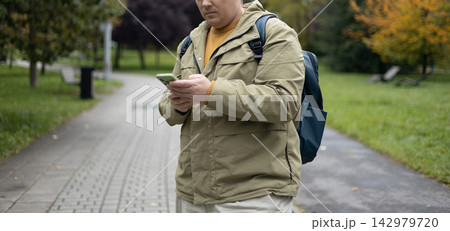 Handsome young 30s man with backpack using mobile phone, texting on smartphone outdoor in park. Technology and communication banner. Autumn time, no face 142979720
